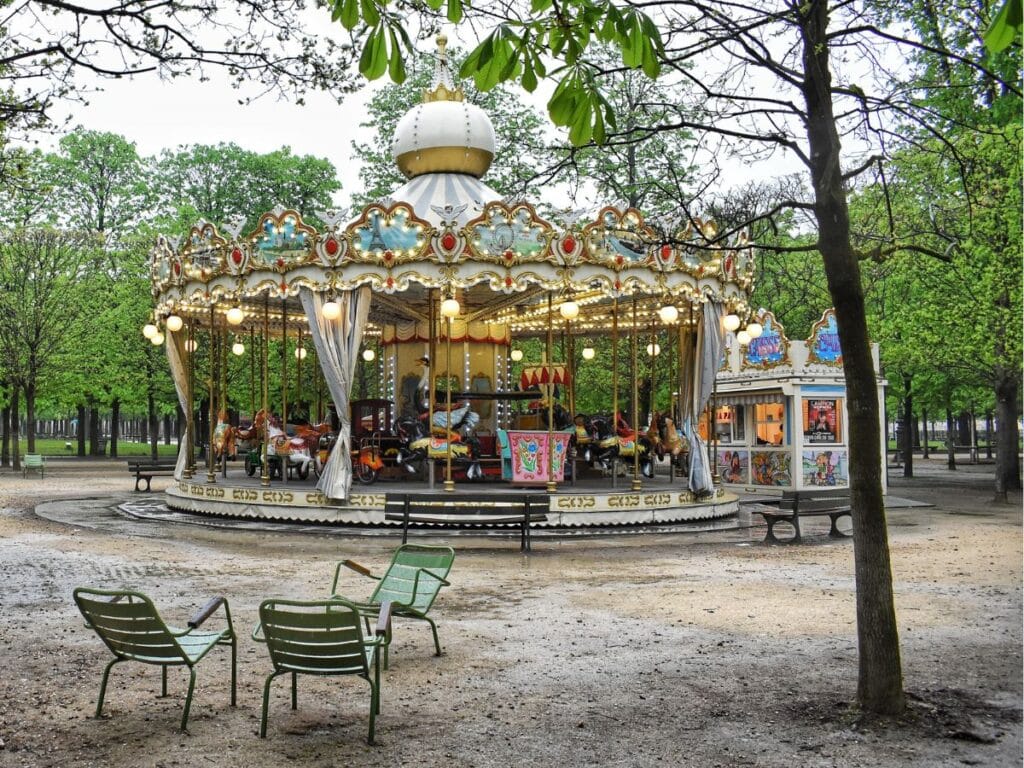 Carousel with vibrant lights in a serene park setting with empty chairs and lush trees.