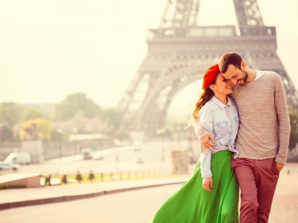 A joyful couple embracing, with the woman in a green skirt and red beret and the man in a casual grey sweater, both smiling in front of the Eiffel Tower on a sunny day.
