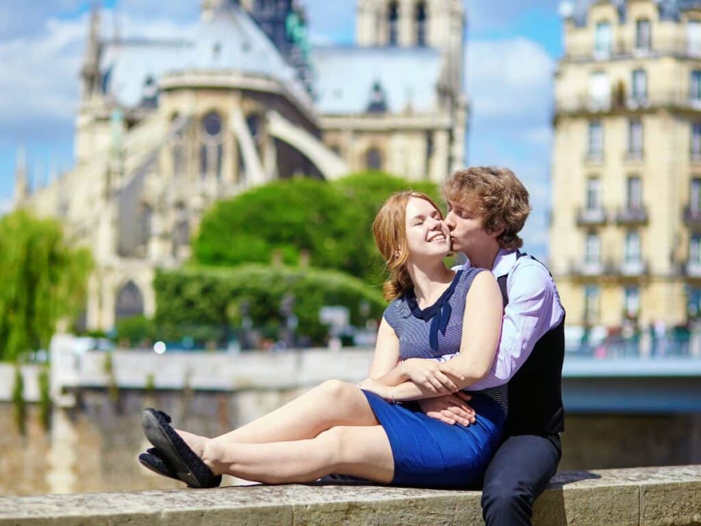 A couple enjoys a sunny day by the Seine with Notre Dame Cathedral in the background; the woman sits on the ledge as the man leans in, both appearing relaxed and happy.