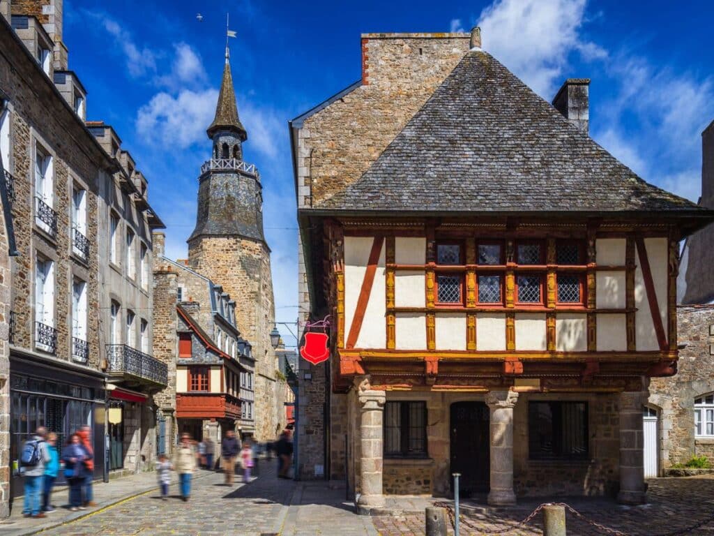 La Maison de la Harpe, an iconic medieval half-timbered house in Dinan, Brittany, with a stone base and intricate wooden framing, standing in a cobbled street with vibrant shops in the background.