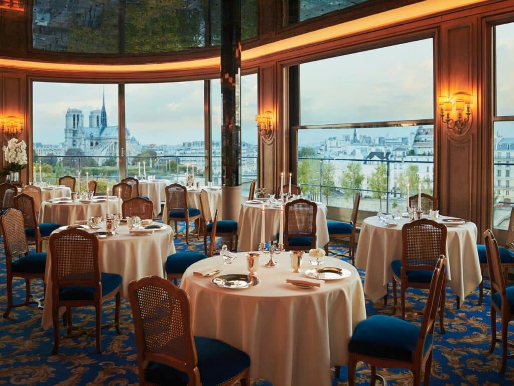 Elegant restaurant interior with tables set for fine dining, featuring blue velvet chairs, crisp white tablecloths, and large windows offering a stunning view of Notre-Dame Cathedral in Paris.