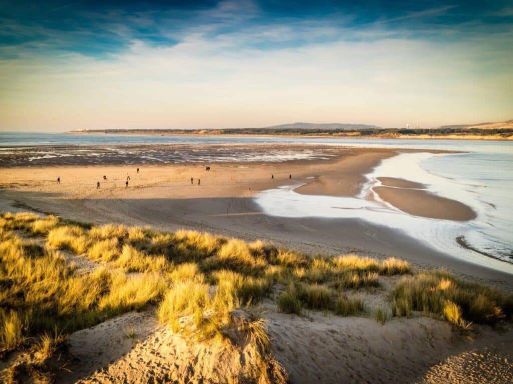 Golden hour at a tranquil beach with people and dunes under a soft sky.