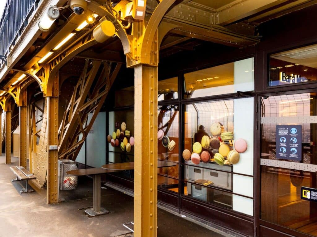 GPT

Inside the Eiffel Tower, a cozy macaron bar invites visitors with a display of colorful macarons arranged in the window. The iconic ironwork of the tower looms overhead, warmly lit by a series of lights, and COVID-19 health advisory signs are visible, indicating the ongoing safety measures.