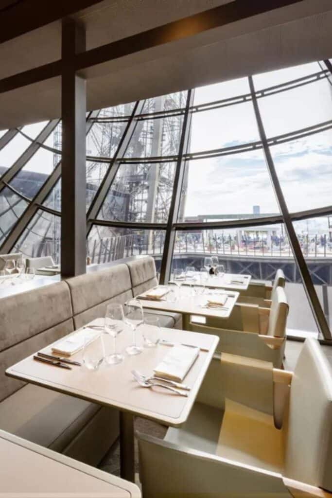 Elegant dining setup inside Madame Brasserie restaurant, with cream-colored booths and tables, pristine table settings, and large glass windows showcasing the intricate metalwork of the Eiffel Tower