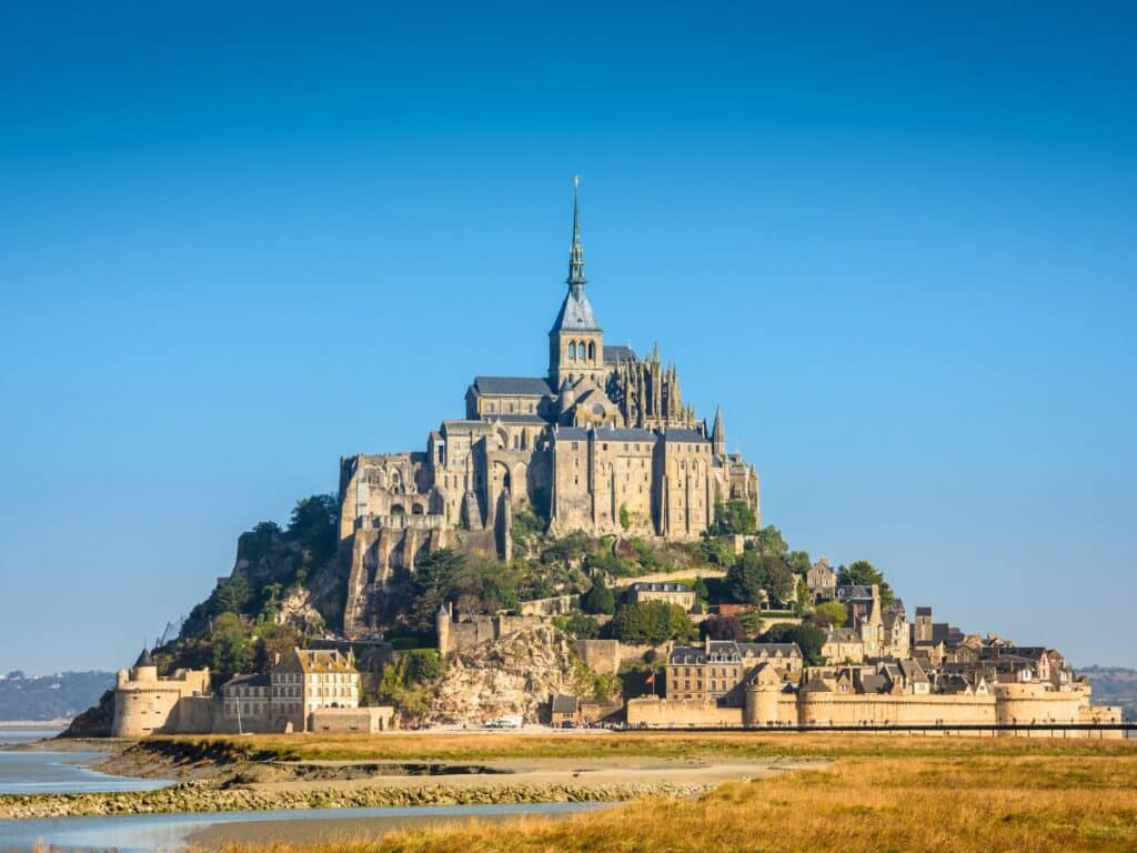 Mont Saint-Michel in Normandy, France, capturing its majestic silhouette against a clear blue sky. This iconic island commune features medieval architecture with the abbey perched atop, surrounded by low tides on the sandy shore.