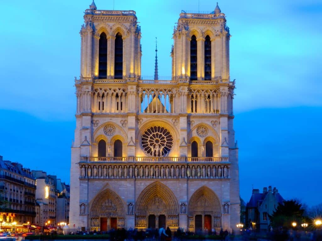 Twilight view of Notre-Dame de Paris with its Gothic façade and twin bell towers illuminated, as seen from the bustling streets filled with the warm glow of street lamps and city life.