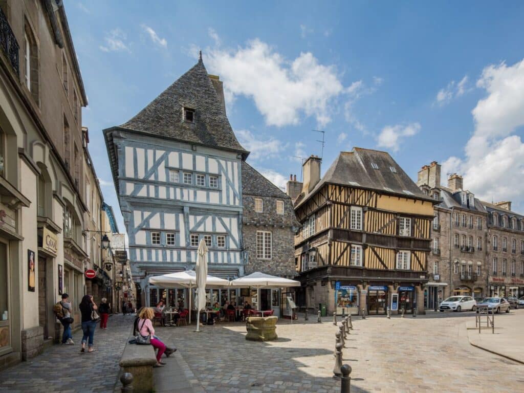 Place des Merciers et des Cordeliers in Dinan, a bustling square surrounded by medieval half-timbered houses, a mix of blue and brown wooden beams, and outdoor cafes, capturing the essence of local life.