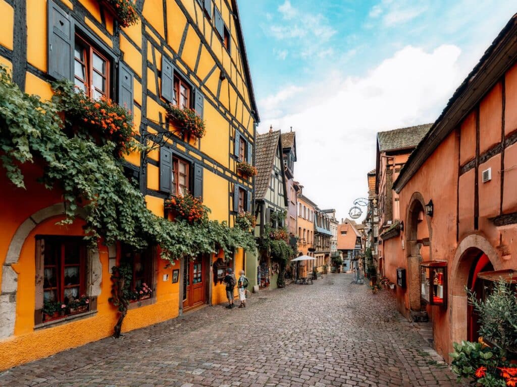 Cobblestone street in a quaint European town with traditional half-timbered houses in bright yellow and orange, adorned with lush green plants and flowers, under a cloudy sky.