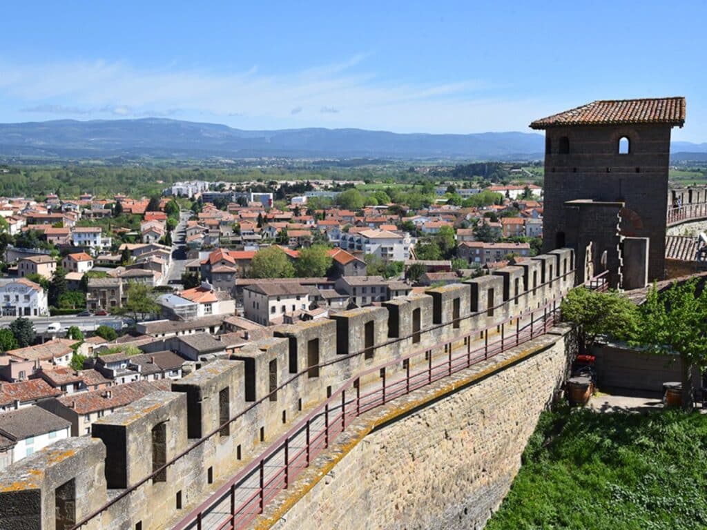 View from the ramparts of Carcassonne Castle showing the crenellated walls and a watchtower, with the modern town and rolling hills in the background, illustrating the historical blend of royal and military presence in the region.