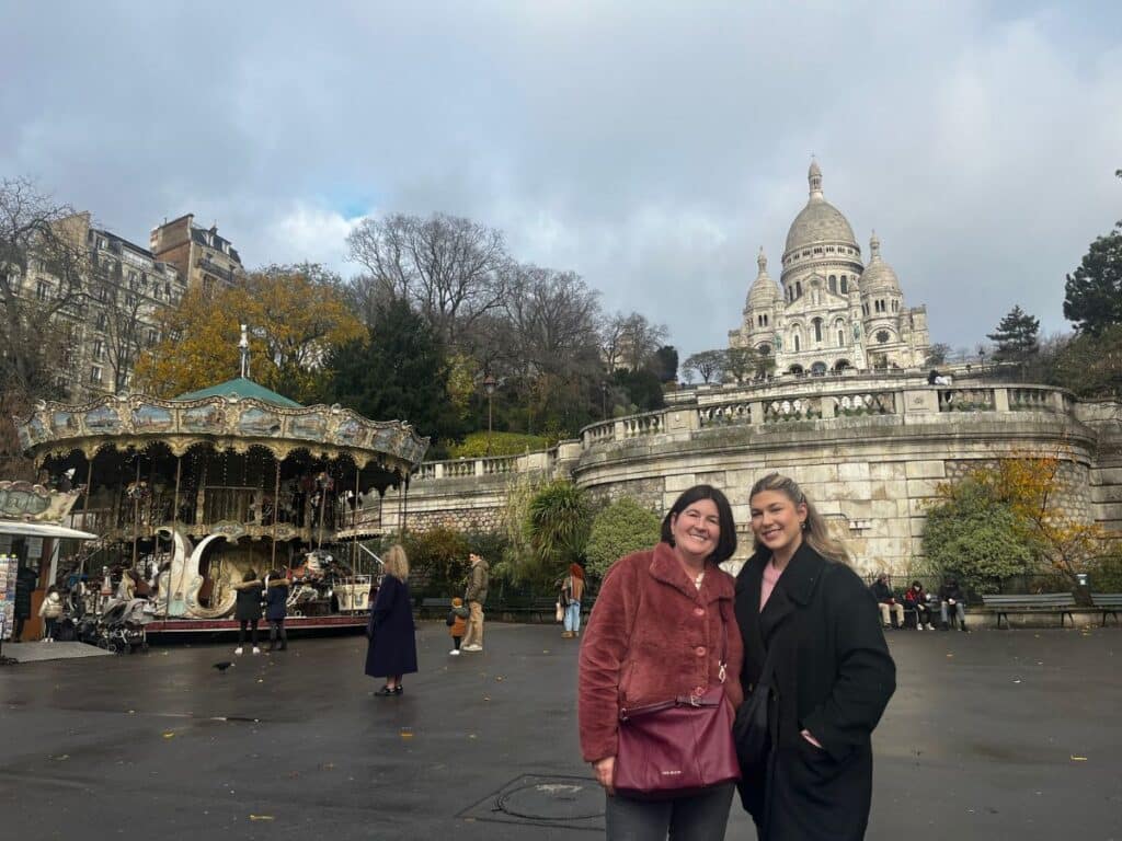 Two smiling women in front of a carousel with the Sacré-Cœur in the distance.