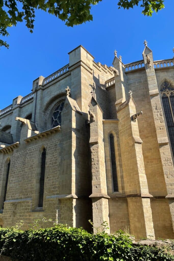 Side exterior view of Saint Michael's Cathedral of Carcassonne with its Gothic architectural elements, flying buttresses, and statues, framed by blue skies and green foliage