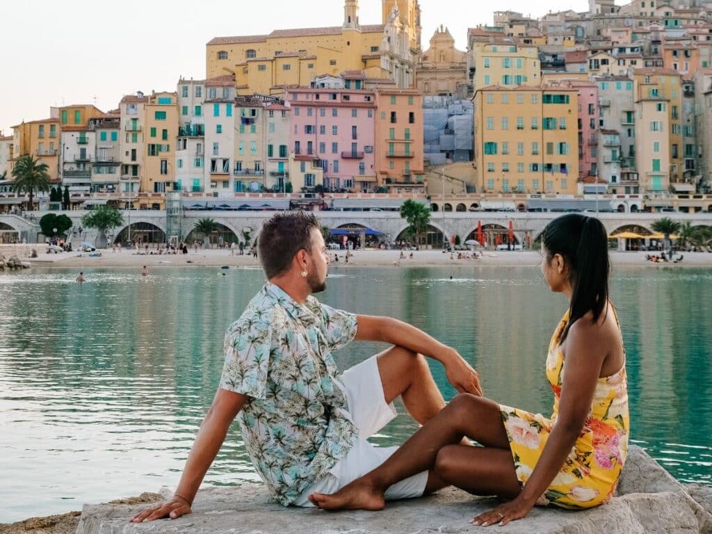 A man and woman sit facing each other on a rock, with a picturesque view of colorful buildings by the sea, likely in a Mediterranean coastal town, sharing a quiet moment during golden hour.