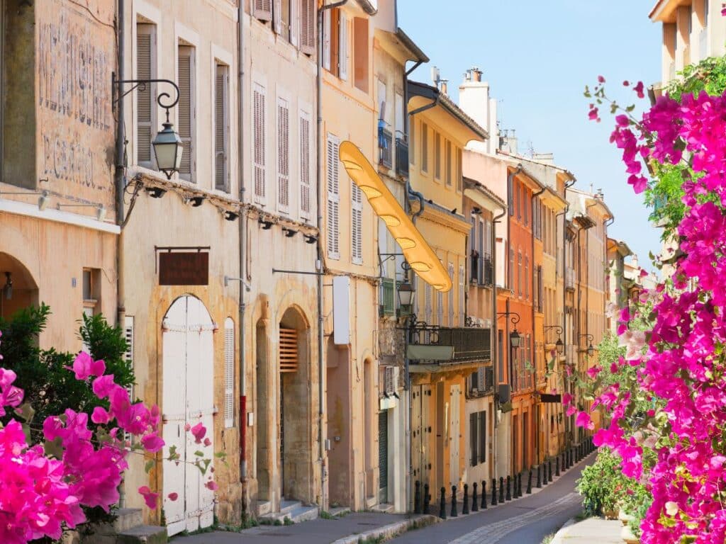 Sunlit French street lined with colorful old buildings and vibrant pink bougainvillea.