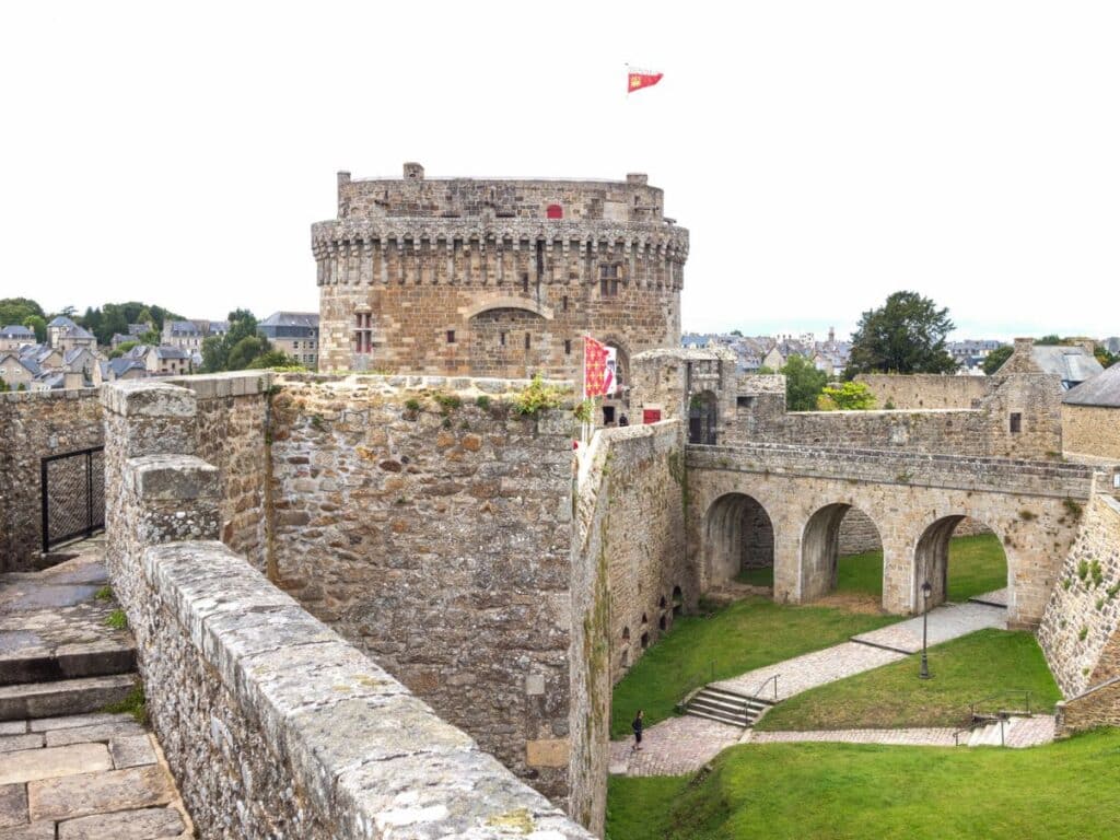 The Castle at Dinan, a fortified medieval stone fortress with a large round tower and ramparts, overlooking the town and surrounded by a green moat, symbolizing the historical significance of Dinan.
