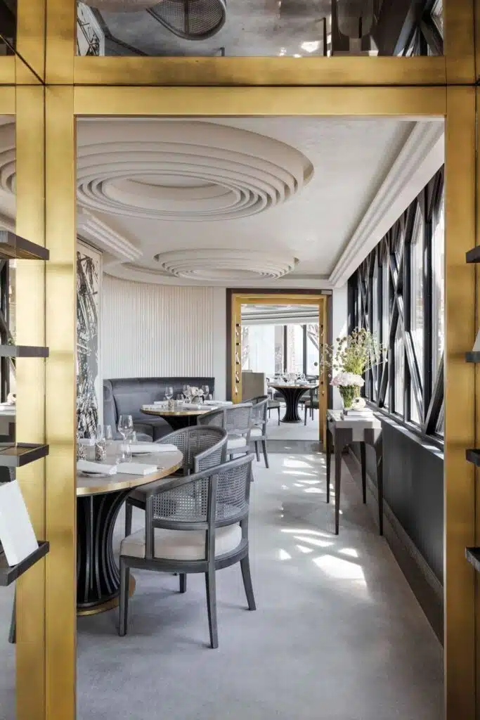 The opulent interior of Le Jules Verne restaurant, featuring golden trimmings, sculptural white ceiling details, and elegant dining arrangements, with natural light streaming through geometric window frames.