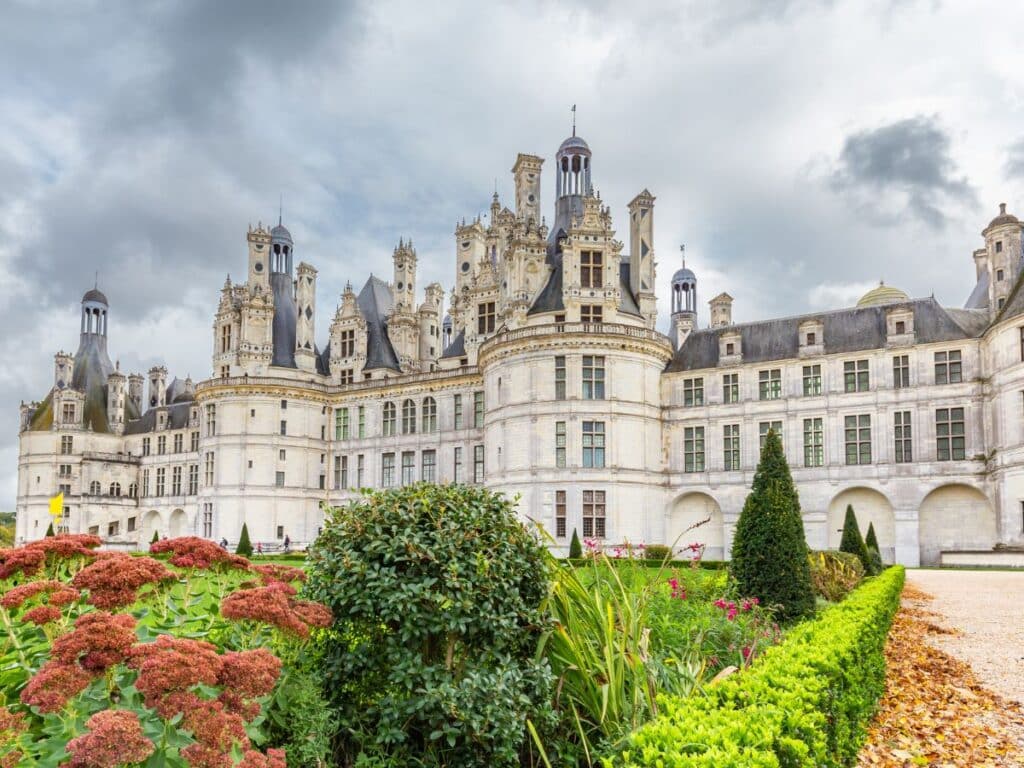 Majestic stone chateau with ornate towers under a cloudy sky, flanked by vibrant garden beds.