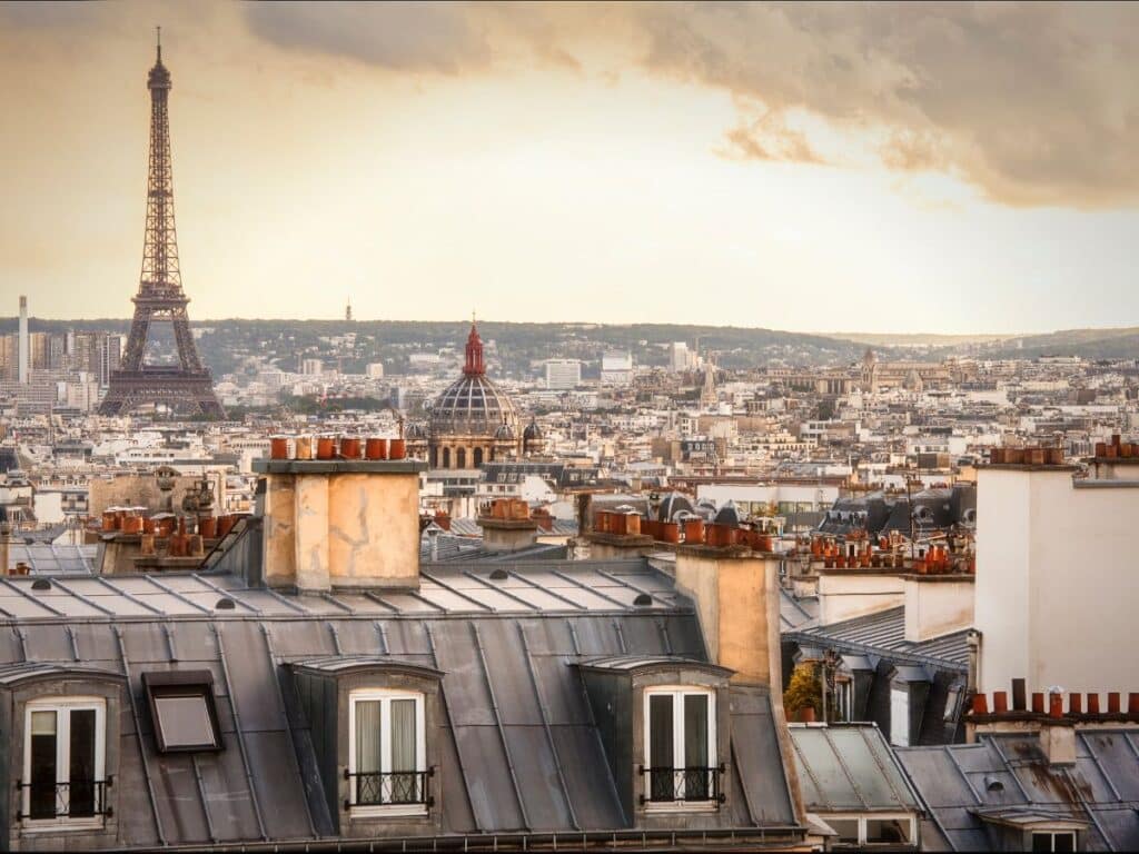 Panoramic view of Paris rooftops at sunset with the iconic Eiffel Tower in the distance. Foreground shows classic Parisian buildings with zinc roofs and chimney pots, set against a softly lit sky.