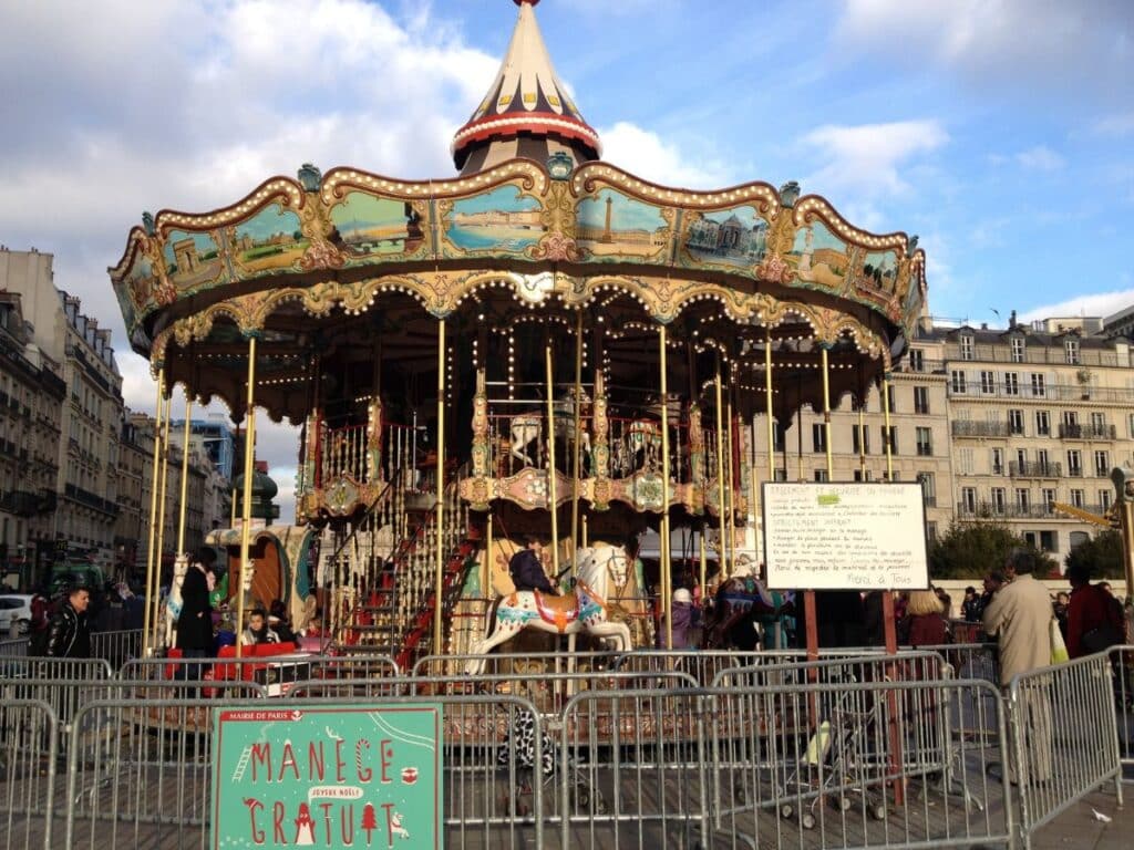 Busy Parisian carousel enclosed by fences, with a sign reading 'MANÈGE GRATUIT'.