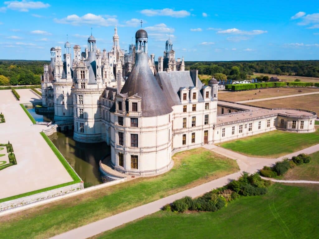 Aerial view of the Château de Chambord in France, displaying its intricate turrets and white stone façades surrounded by a moat, with expansive green lawns and forested landscape stretching into the distance.
