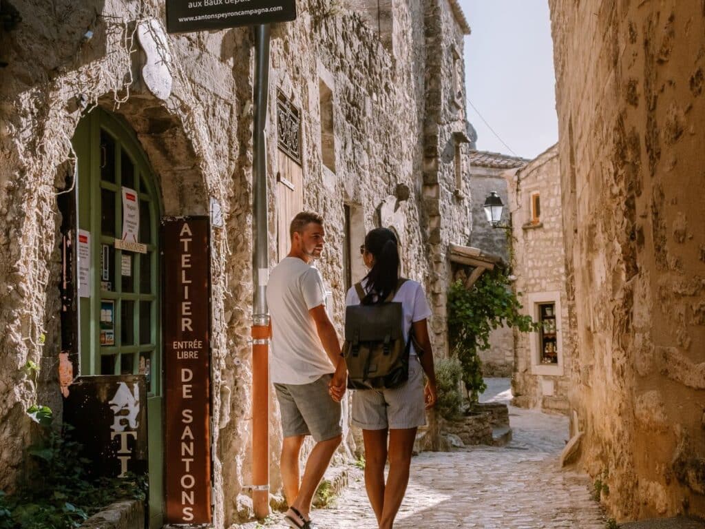     A couple walks hand in hand down a narrow, cobblestoned street in a rustic village, passing by a shop with a sign that reads "Atelier de Santons," indicating a crafts workshop, enjoying the charm of the old town.