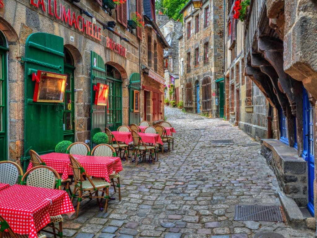 A cobblestone street in Dinan, Brittany, with a row of vibrant green-painted restaurants and cafés, outdoor seating covered with red-checkered tablecloths, inviting visitors to enjoy the local cuisine.