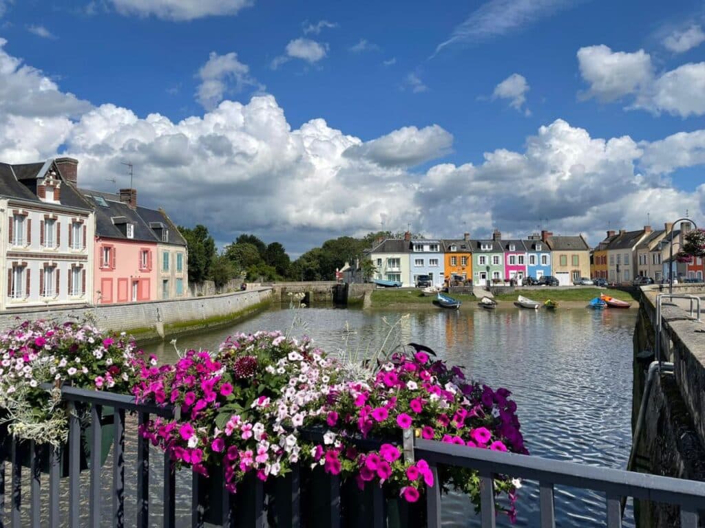 Scenic view of a calm canal in France lined with colorful houses in shades of pink, blue, and pastel, viewed from a flower-adorned bridge under a dramatic sky with fluffy clouds.