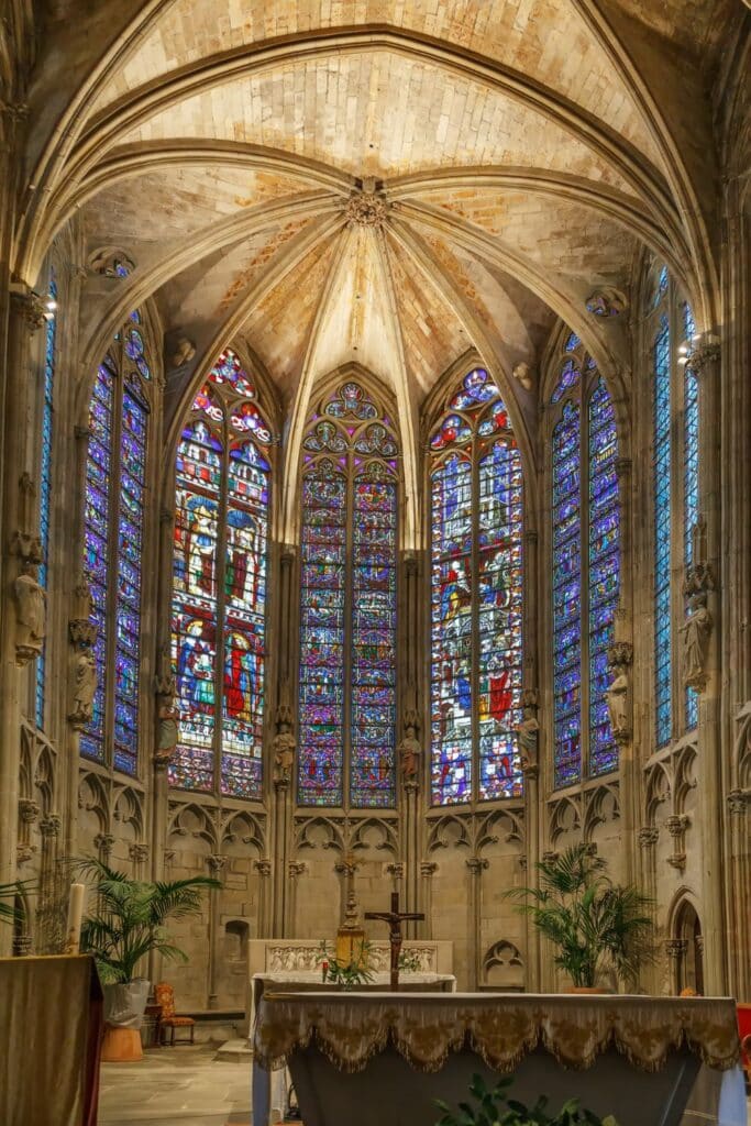 Interior of the Carcassonne Cathedral, featuring a vaulted ceiling and tall stained glass windows that illuminate the altar area with colorful light.