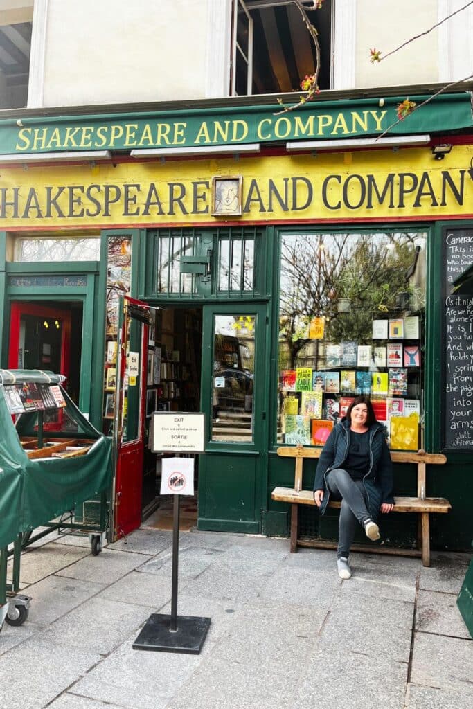 Woman seated on a bench outside a colorful bookshop with the sign 'Shakespeare and Company'.