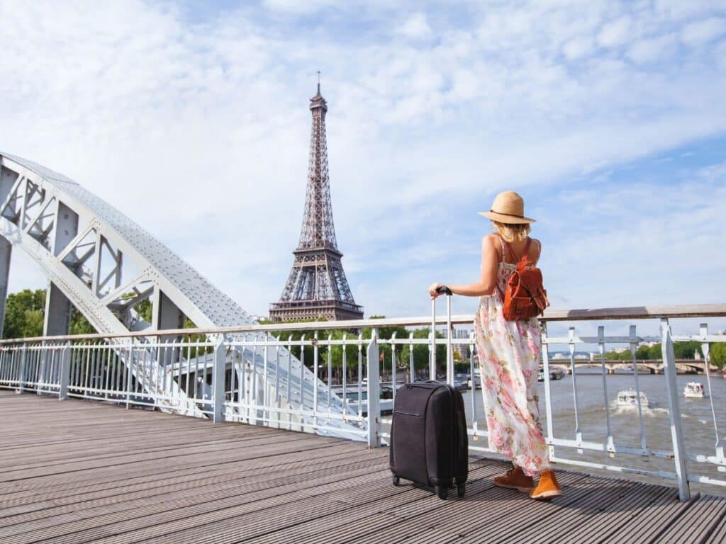 Woman with suitcase on Paris bridge, Eiffel Tower in background, sunny day.