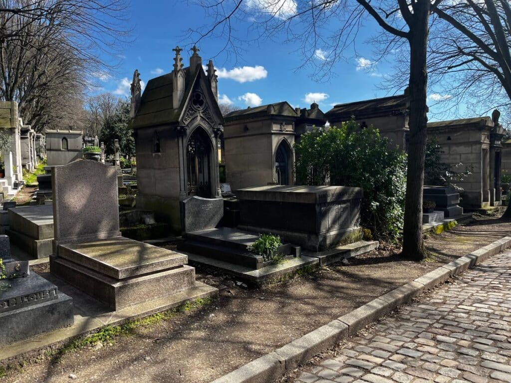 A serene cemetery with Gothic-style mausoleums and bare trees under a blue sky. Keywords: cemetery, Gothic mausoleums, serene, headstones, blue sky