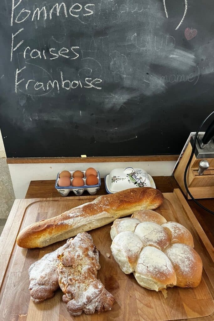 Fresh bread on a cutting board with eggs and a chalkboard listing in French.