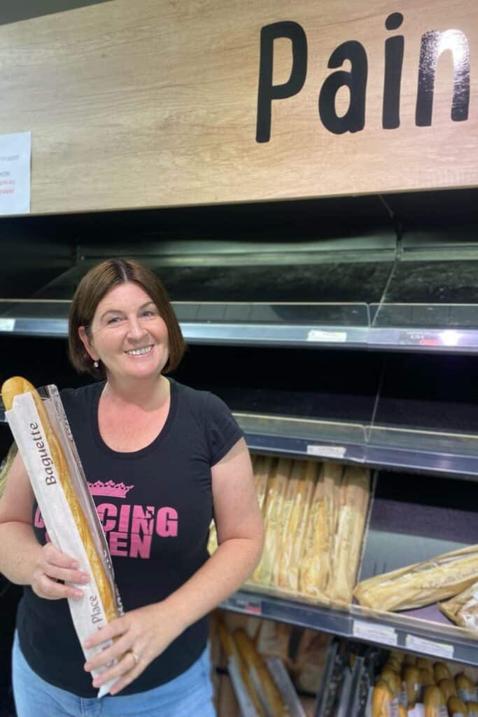 Woman holding a baguette in a bakery under a 'Pain' sign.