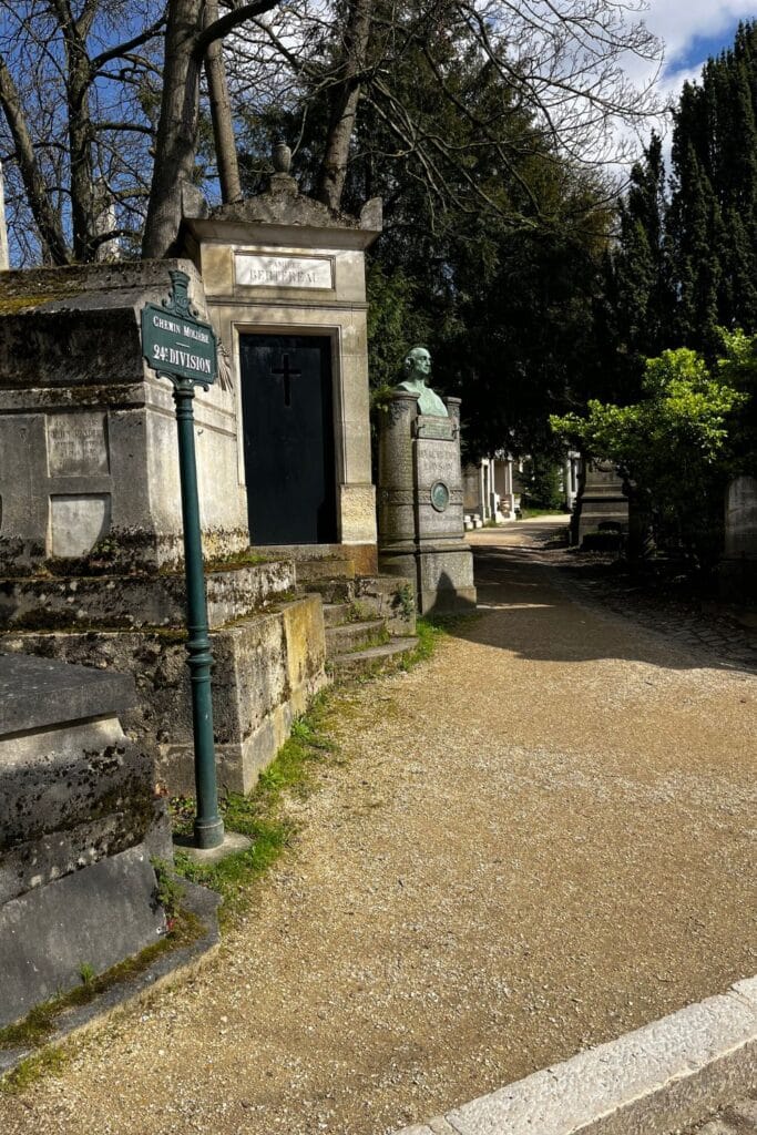 Shadow-dappled path with a bust statue beside a tomb under a clear sky.