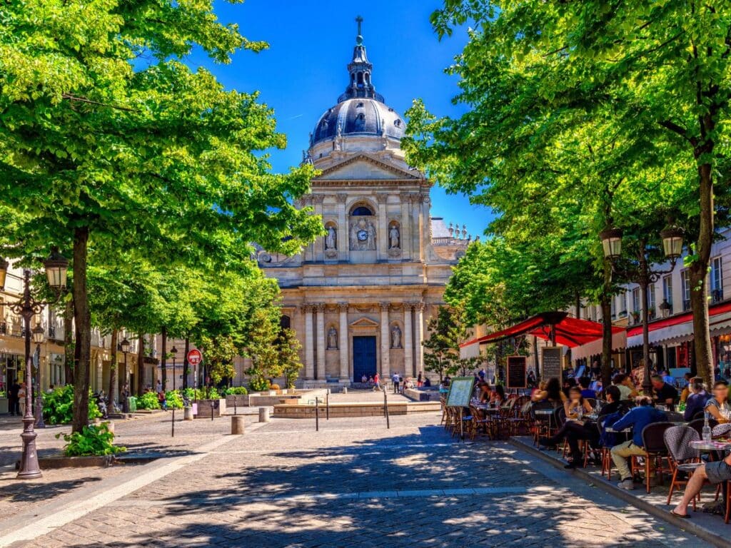 Sunny Parisian square with lush trees and café patrons facing a historic stone building with a dome.