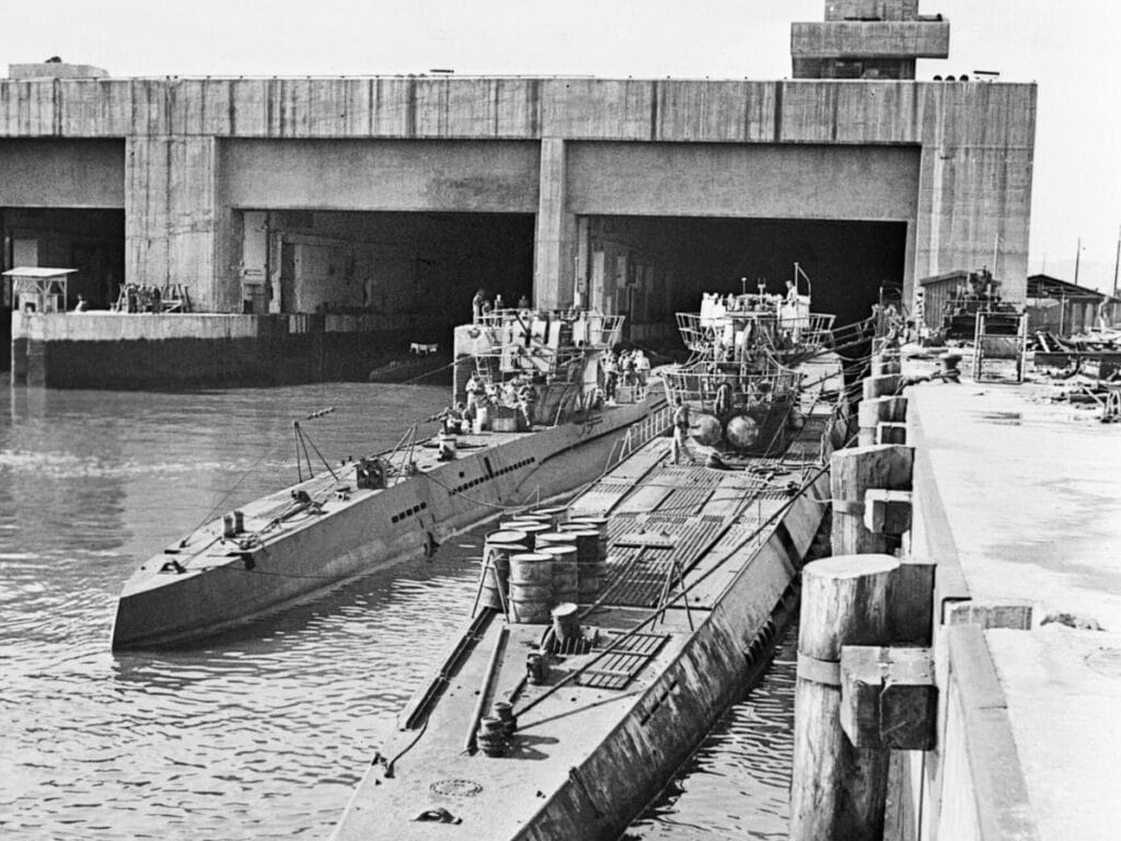 Historic black and white photo showing a German U-Boat from World War II docked at a submarine pen in La Rochelle. The massive concrete structure overshadows the submarine and various naval activities can be seen in the background, capturing a moment of maritime military history.