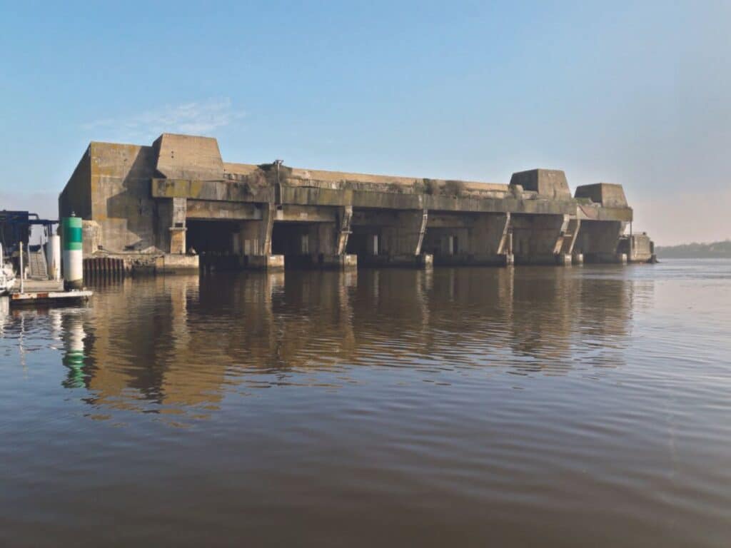Photograph of the imposing World War II bunkers at La Rochelle, partially submerged in the calm waters. The concrete structures stand as silent remnants of history, their stark, weathered exteriors reflecting in the water, juxtaposed against a peaceful, hazy sky.