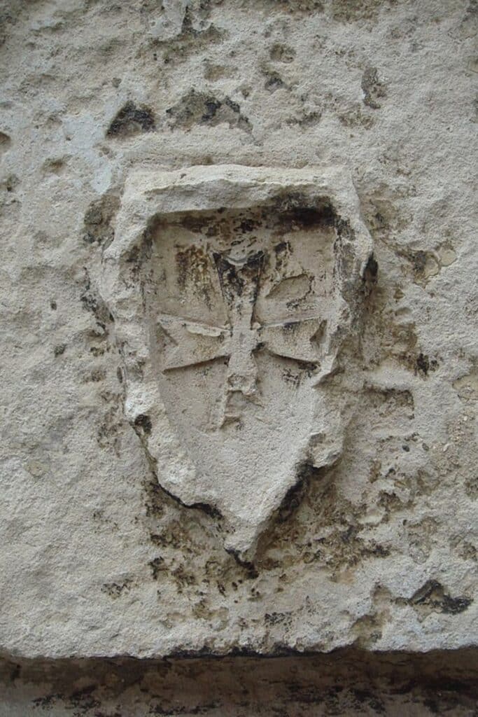 A weathered stone carving of the Templar Cross, subtly etched into the facade of an ancient wall in La Rochelle. The emblem, known for its association with the Knights Templar, shows signs of erosion but retains its distinct symmetric shape and design, symbolizing the history of La Rochelle France.