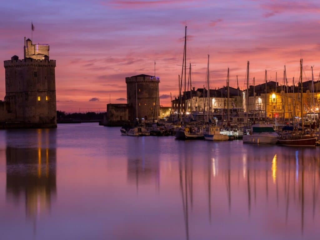 Twilight descends over the historic port of La Rochelle, with the medieval towers of the Vieux-Port standing sentinel. Sailing boats are moored in the calm harbor waters, reflecting the pink and purple hues of the sunset sky. The city's warm lights begin to twinkle against the evening backdrop, creating a serene and picturesque scene.
