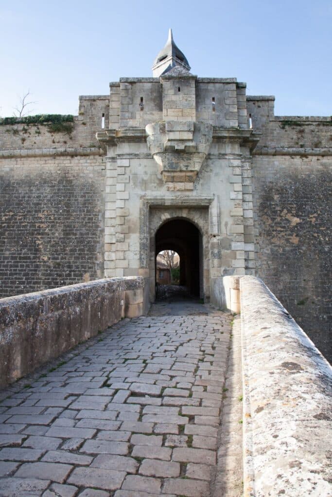 Blaye Citadel in the Gironde, featuring a medieval stone gateway with a distinctive pointed turret. The structure is constructed from aged stone, displaying wear that hints at its historical significance. A cobbled pathway leads through the archway, inviting exploration into the fortress that has stood guard over the region for centuries. The clear sky and natural light enhance the texture and detail of the ancient masonry.