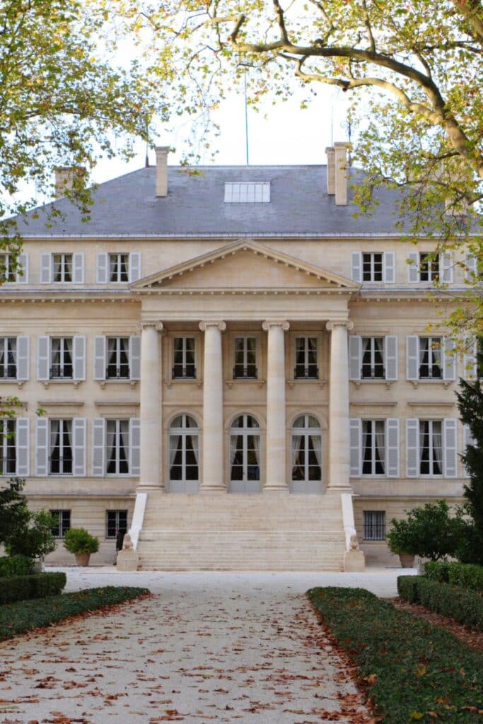 Château Margaux, a renowned wine estate, captured from its front gravel path lined with neatly trimmed hedges. The classic French architecture is highlighted by elegant columns and multiple windows with shutters, set against a backdrop of mature trees with autumn leaves. This historic building exudes a sense of tradition and luxury, characteristic of a prestigious wine estate.