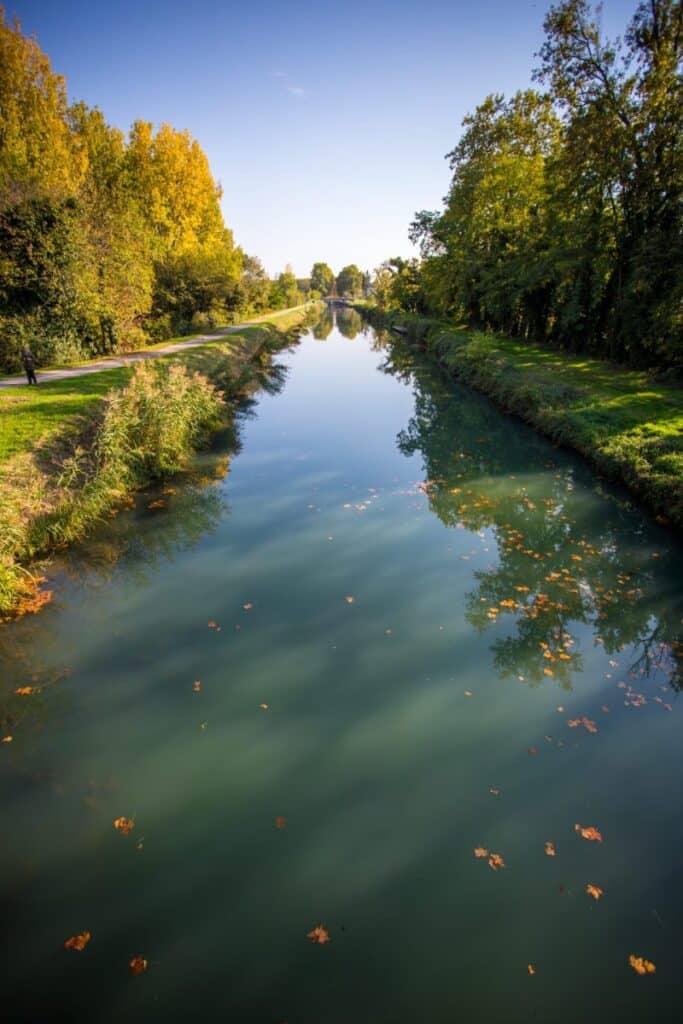 A tranquil canal scene in the Entre Deux Mers region, flanked by lush, colorful autumn trees reflected in the calm, clear water. A path runs alongside the canal, where a person is visible walking, enjoying the serene environment. Fallen leaves dot the surface of the water, adding a touch of seasonal charm to the peaceful setting. The scene captures the essence of a peaceful autumn day in a picturesque rural landscape.