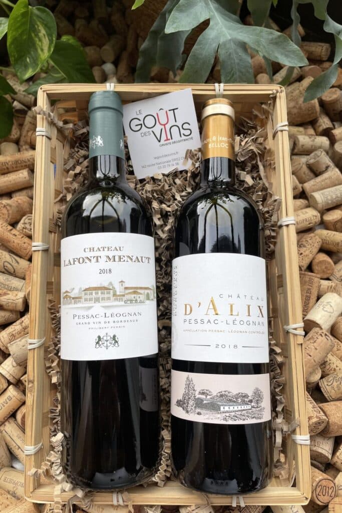 A wooden gift box containing two bottles of Pessac-Léognan wine, one from Château Lafont Menaut and the other from Château d'Alix, both from the 2018 vintage. The bottles are nestled in straw, with a business card reading "Le Goût des Vins" placed in between them. Behind the box, a backdrop of corks and lush green foliage adds a natural, rustic charm to the scene, emphasizing the theme of wine appreciation.