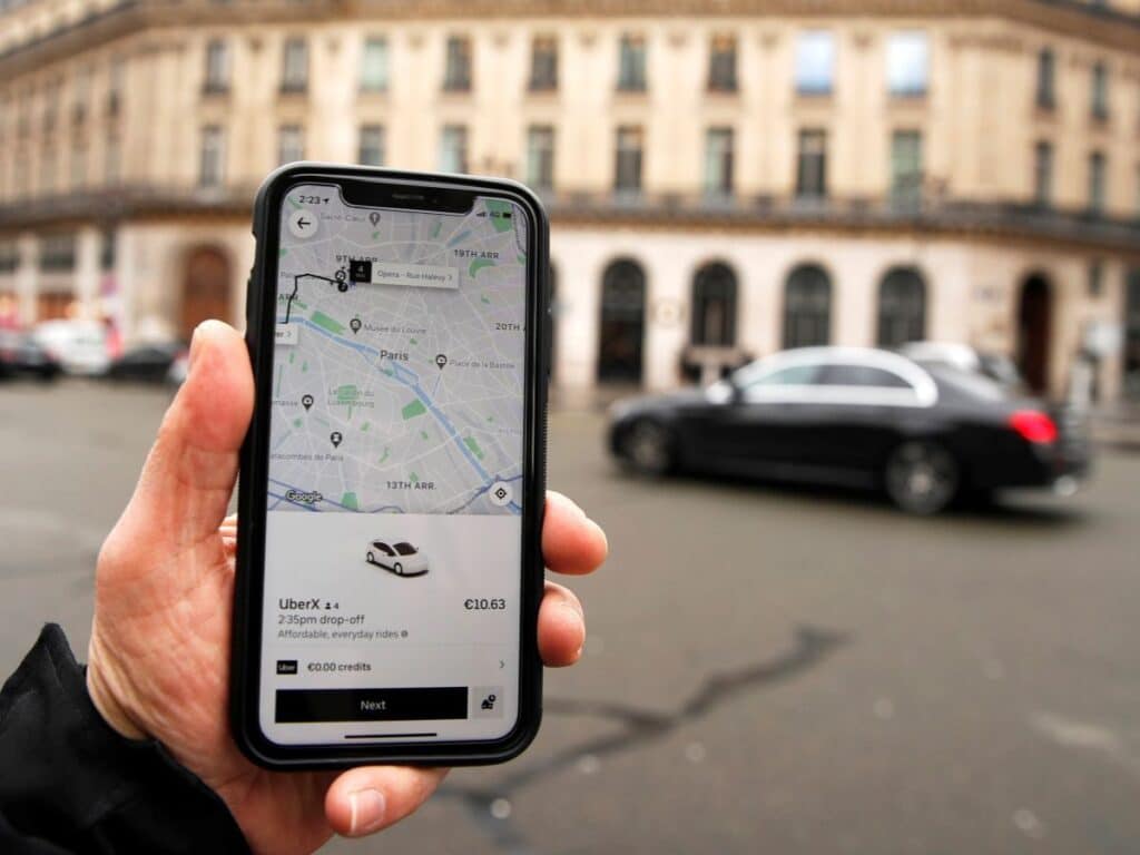 A hand holding a mobile phone with an Uber taxi App loaded and an old Parisian building in the background with cars going past.