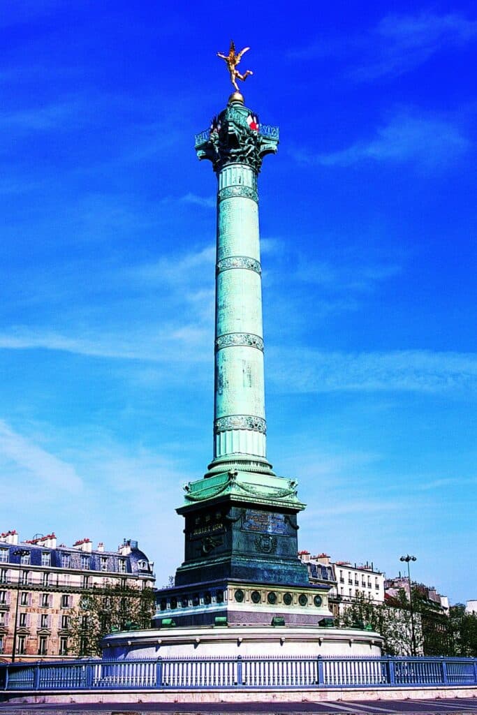The July Column (Colonne de Juillet) located at the Place de la Bastille in the 11th arrondissement of Paris. The column is topped with a golden statue, and the bright blue sky serves as a backdrop. The base of the column includes an inscription and reliefs, with surrounding Parisian buildings in the background.