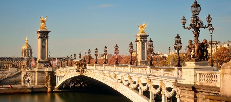 The ornate Pont Alexandre III bridge in Paris, featuring golden statues and intricate lampposts, spans the Seine River. The domed Les Invalides building is visible in the background under a clear blue sky.