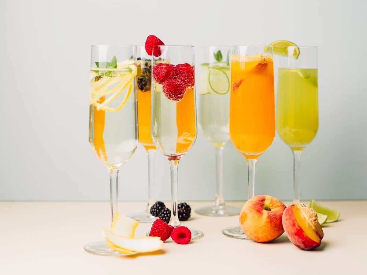 A colorful array of summer cocktails in tall glasses, garnished with fresh fruits and herbs, with additional fruits placed around the glasses on a light surface.