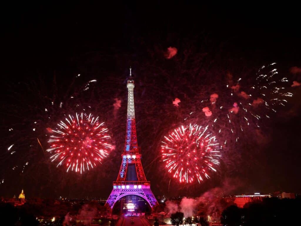 The Eiffel Tower illuminated with red, white, and blue lights, surrounded by vibrant red fireworks during a night celebration in Paris.