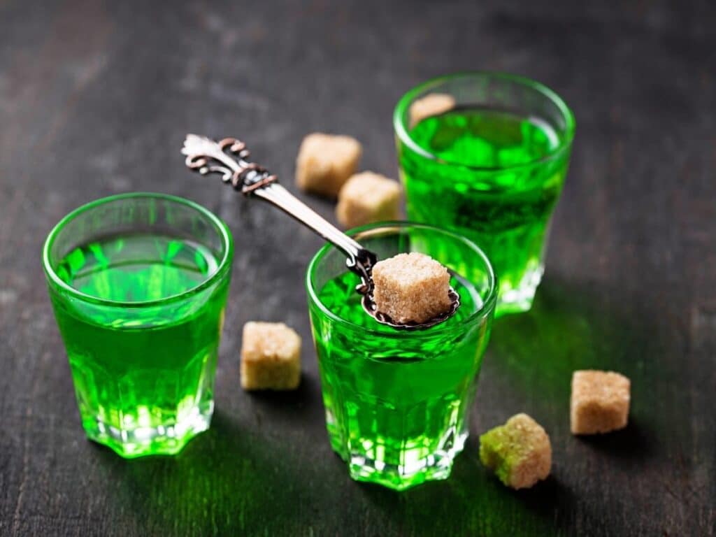 Three green absinthe drinks in ornate glasses, with sugar cubes placed on a traditional absinthe spoon, set against a dark background.