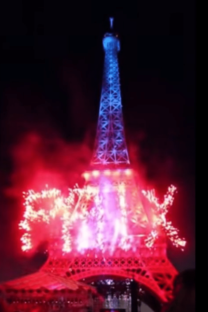 The Eiffel Tower illuminated with red, white, and blue lights, with the word 'PAIX' spelled out in fireworks at the base during a night celebration in Paris.