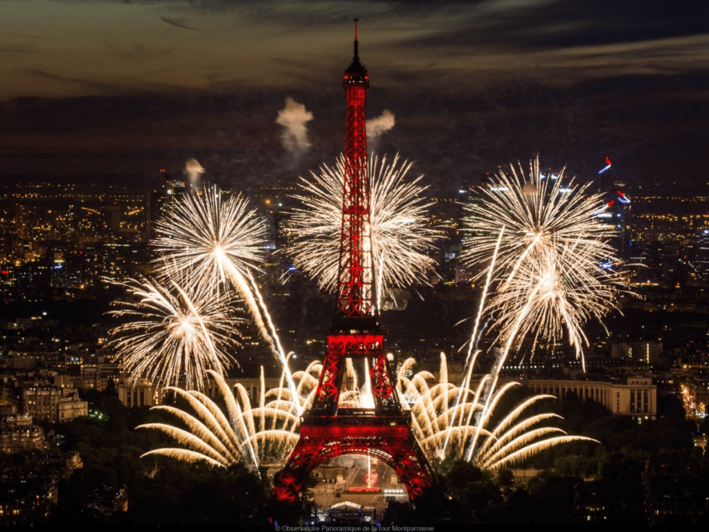 The Eiffel Tower glowing red, surrounded by golden and white fireworks, illuminating the night sky over Paris with city lights in the background.